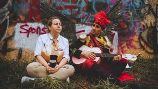 A photograph of two people in costume at a Disco Elysium LARP. Both are sitting cross-legged on the ground, with one holding a cup of coffee while the other strums a guitar wearing a pair of black wings.